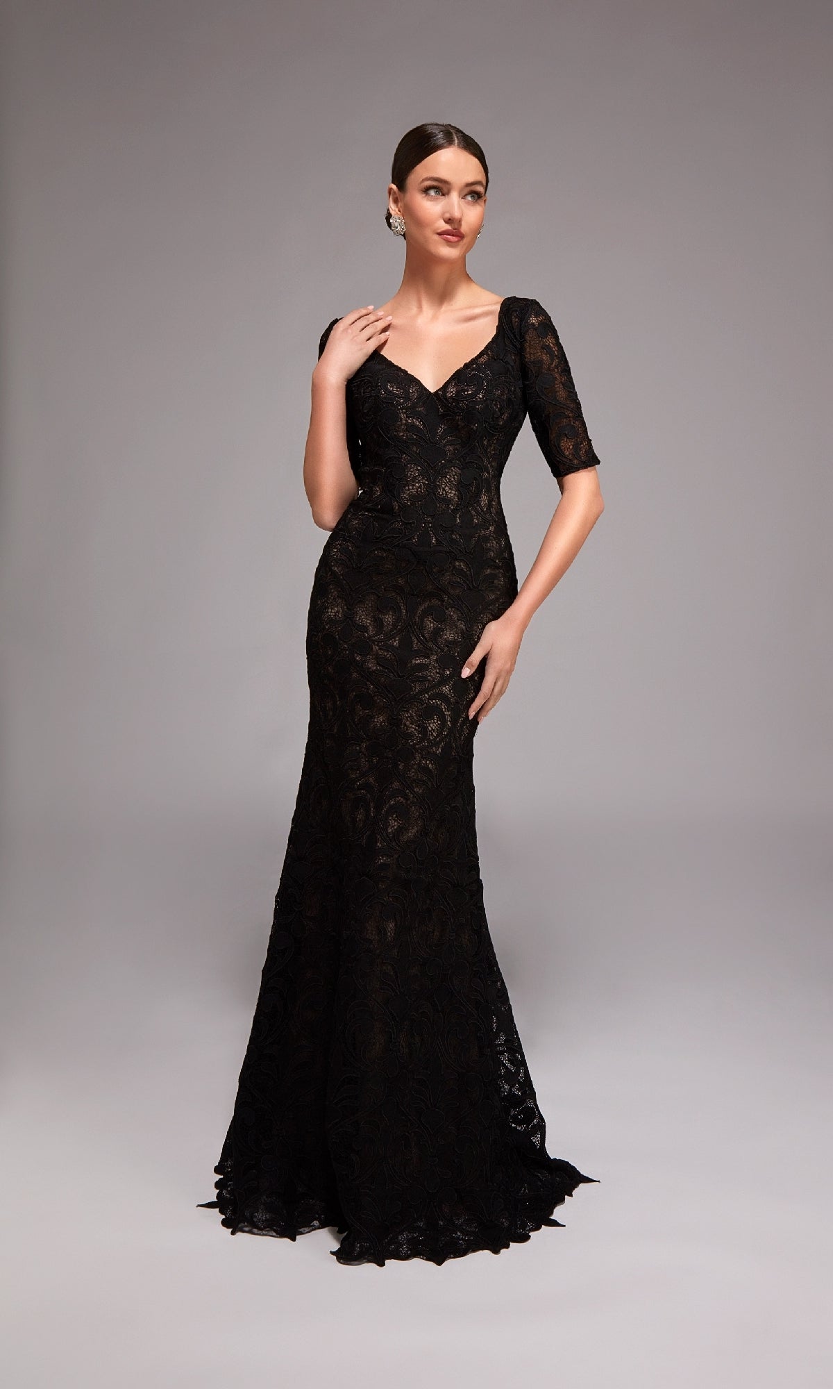 A woman stands against a plain gray background wearing the Alyce 27871 Long Formal Dress, a floor-length, fitted lace overlay gown with short sleeves and a V-neckline.